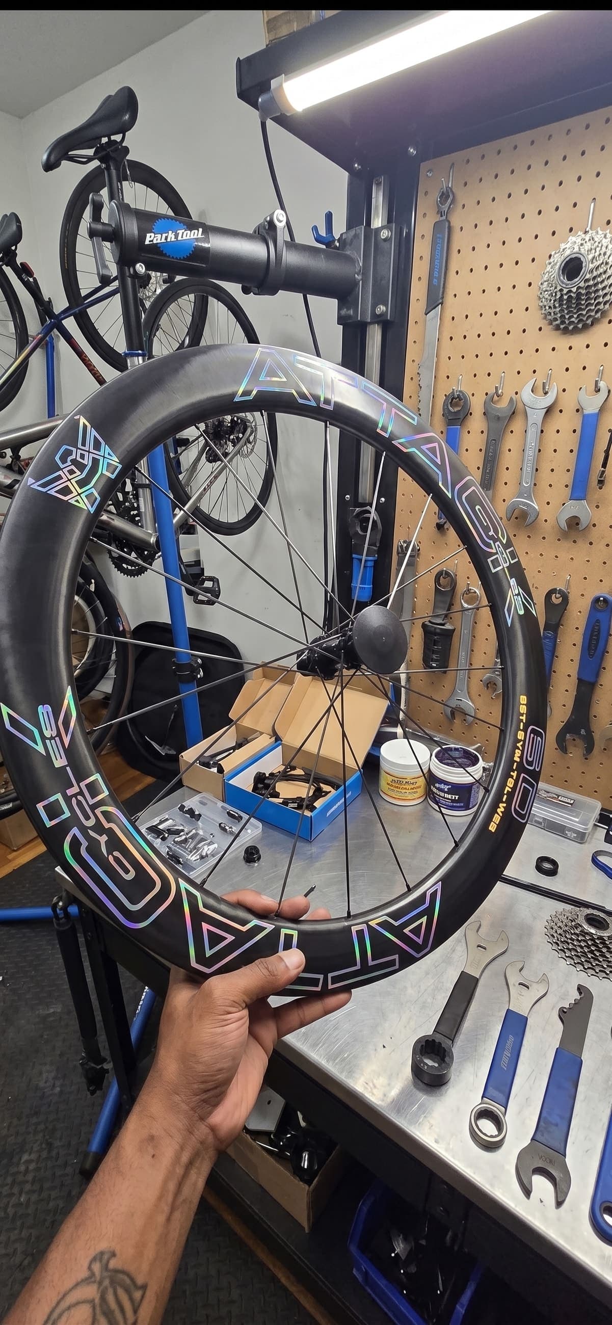 A wheel held at the workbench beside a Park Tool truing stand, wrenches and cassettes on the pegboard behind.